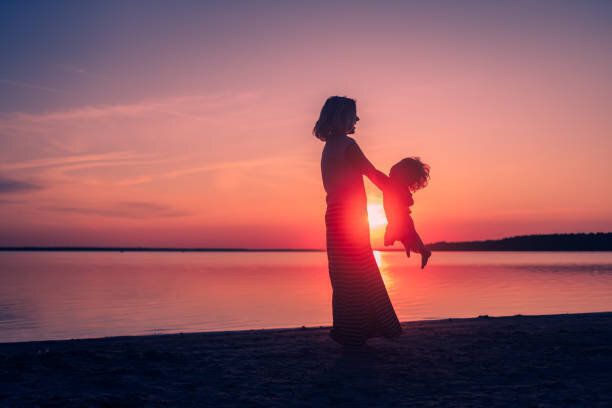 幸せな家族の瞬間。夕焼けの背景に赤ちゃんを回転させるお母さんのシルエット。美しい風景。愛と母性。自由。幼年。姉妹関係。国際女性デー。海の旅