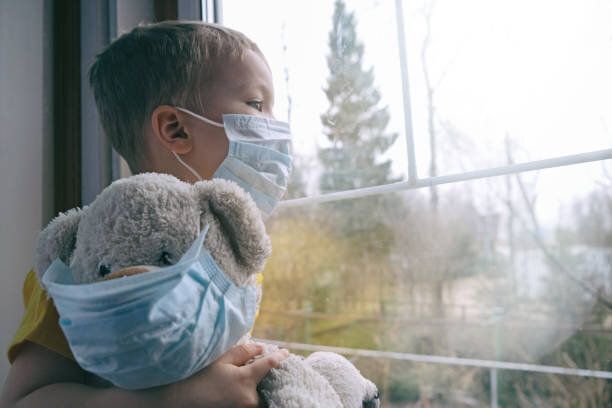 自宅検疫で悲しい病気の子供。保護医療マスクを着用した少年とテディベアは、窓辺に座り、窓の外を見ます。ウイルス保護、コロナウイルス大流行、予防流行。