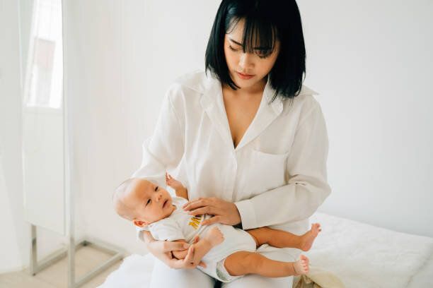 自宅で小さな赤ちゃんを見て自分の幼児を保持白いウェアで陽気な思いやりのある若いアジアの女性の上から