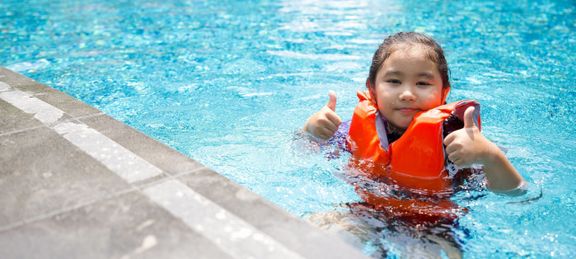 プールで遊ぶアジアの子供。オレンジ色のライフジャケットを着て、親指を立てながら微笑む。