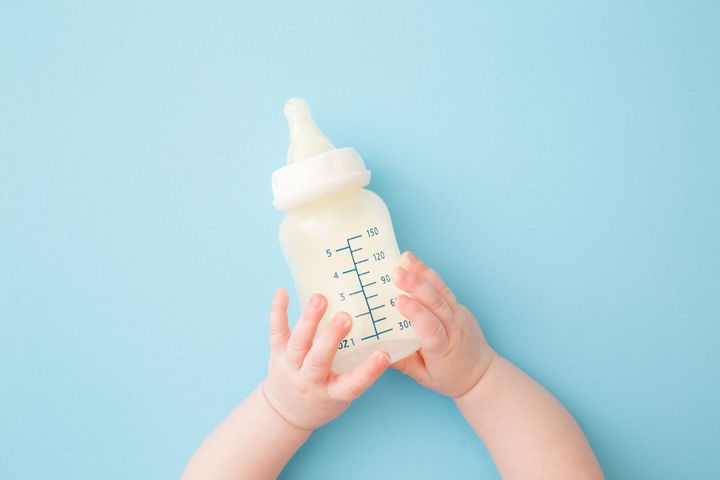 水色の床の背景に牛乳のボトルを保持する幼児の手。給餌時間。パステルカラー。クローズ アップ。視点ショット。上から下のビュー。