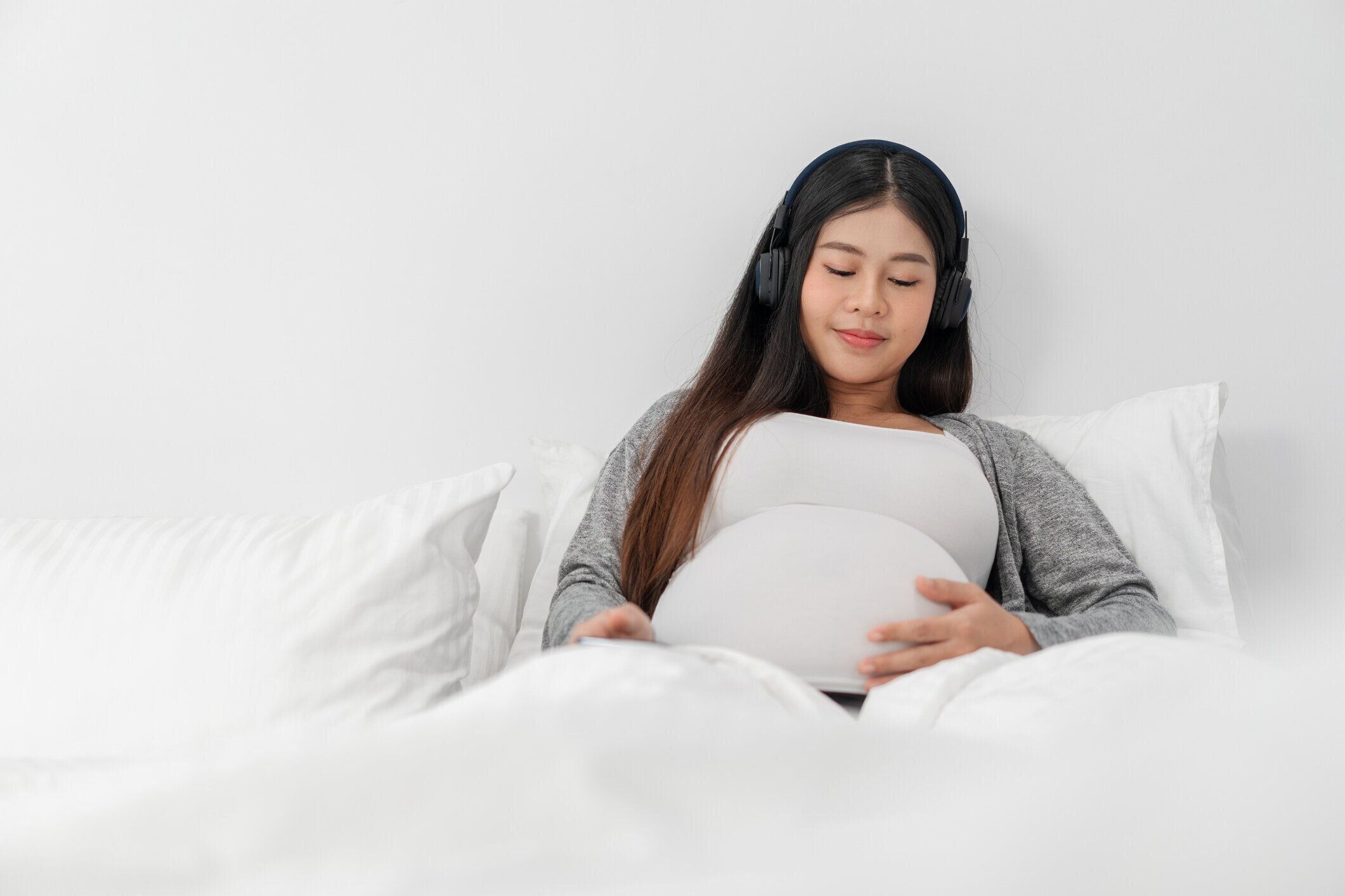 アジアの幸せな妊婦はベッドに座って、彼女の腹に触れて、音楽を聴くためにヘッドフォンを着用しています。妊娠, 母性, 人と期待の概念