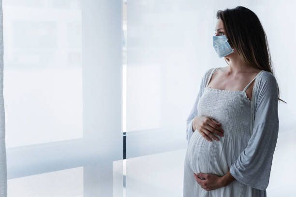 顔の医療マスクを着用した大きな窓のそばに立っている妊婦