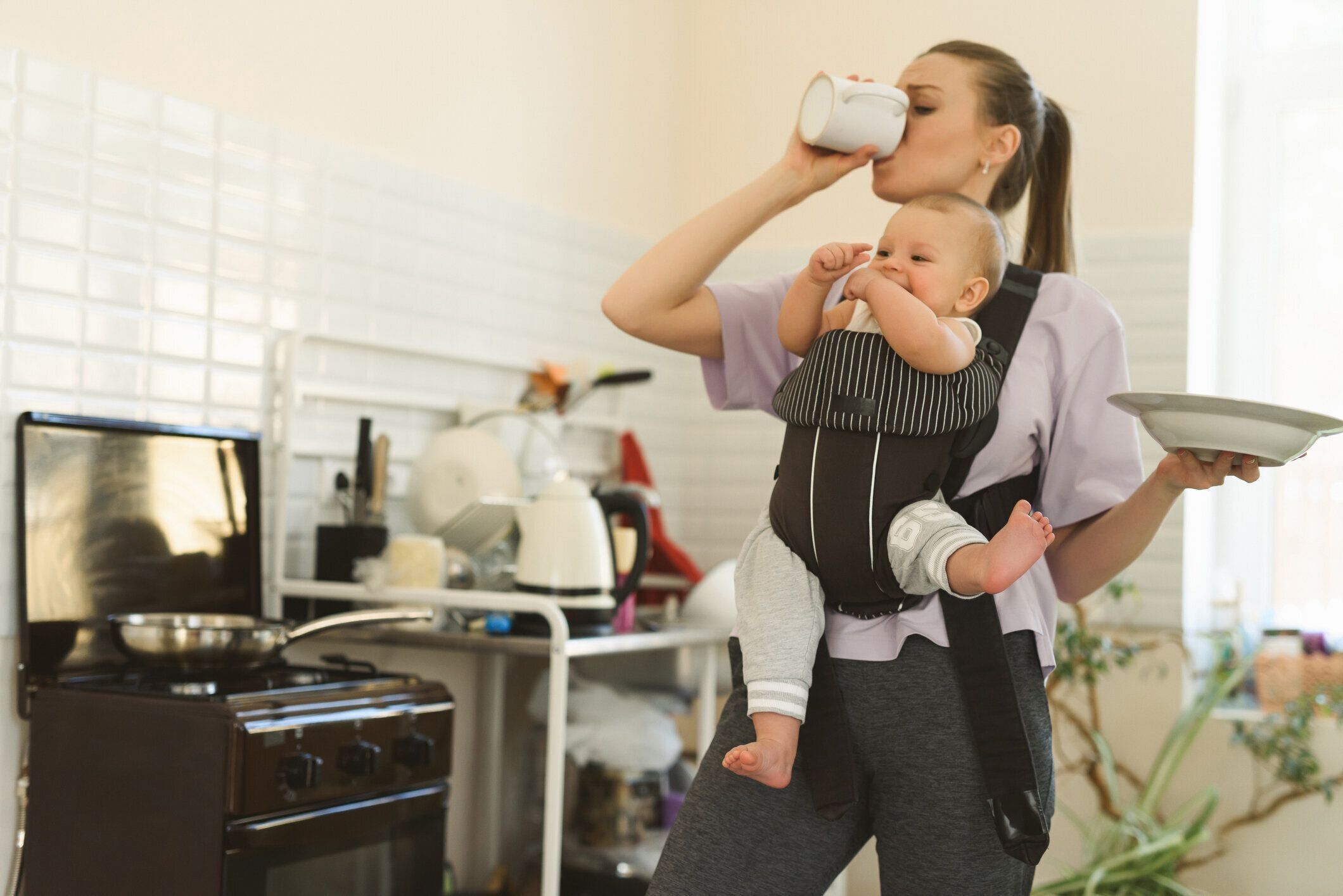 ベビーキャリアで疲れたマルチタスクの母子。子供と台所で若い女の子。母性と孤独の困難