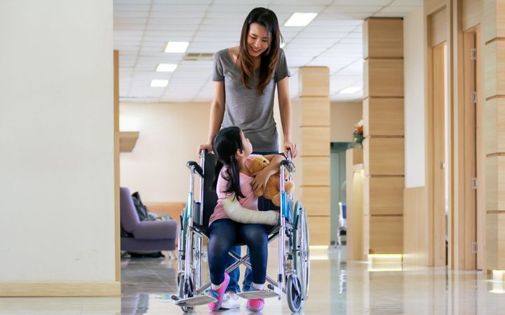 彼女の小さな女の子の娘の世話をしているアジアの若いシングルマザーは、病院の廊下で話している間に怪我をして車椅子に座ります。医療、事故、保険の概念。