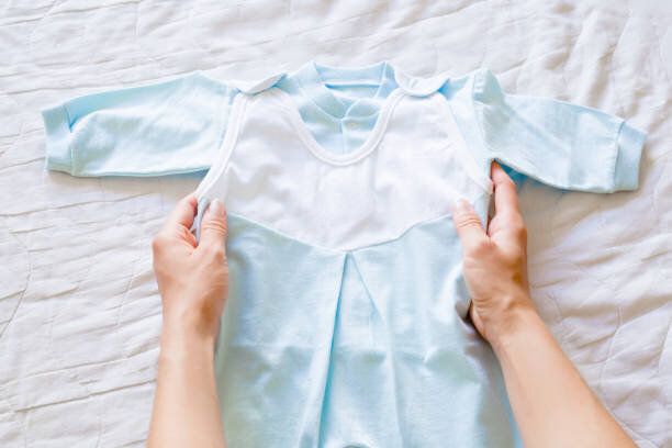 女性の白いベッドのシーツに両手新しい、明るいブルーのボディー スーツ。ベビー服。柔らかな色。