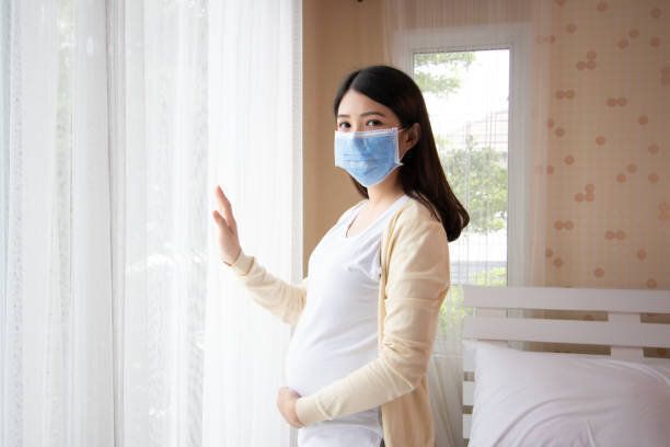 寝室の窓の近くに立って腹に触れる若い美しい妊娠中のアジアの女性。妊娠、ウイルス保護マスク口の概念を身に着けている母性。