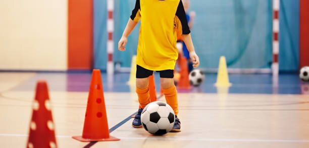 子供用サッカー フットサル トレーニング。サッカー トレーニング ドリブル円錐形をドリルします。屋内サッカー スポーツ ホールにサッカー ボールを持つ若い選手。オレンジ色の制服でプ