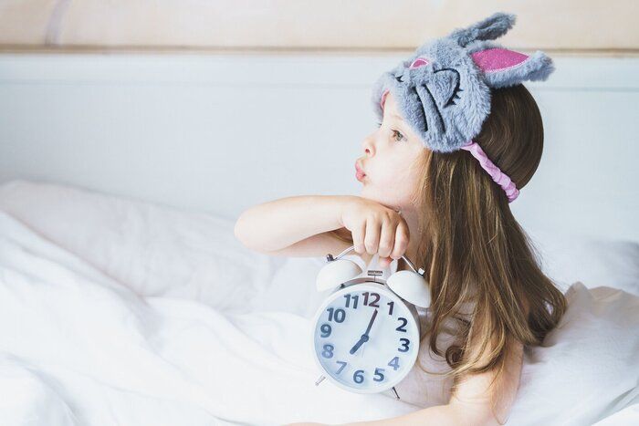 小さな女の子は、彼女の手に目覚まし時計と灰色のバニーぬいぐるみスリープマスクを身に着けている白い毛布の下で朝起きます。子供の睡眠体制の時間。幼稚園への早起き。ママの毎日の�