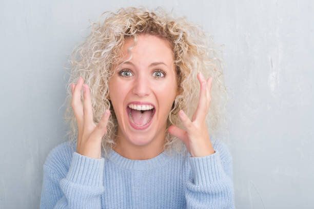 グランジに巻き毛を持つ若いブロンドの女性は祝う狂気の背景をグレーし、驚いて悲鳴上げられたと開いた目が興奮して腕の成功のため。勝者の概念