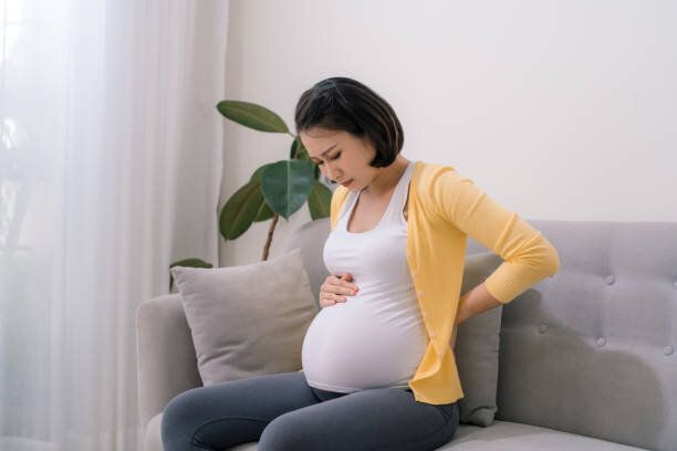 アジアの妊婦は腰痛があり、ソファに座って落ち込んでいる