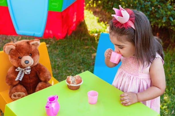 赤ちゃん幼児の女の子がテディベアのテーブルに座っての親友とカップから飲む屋外のティー パーティーで演奏します。