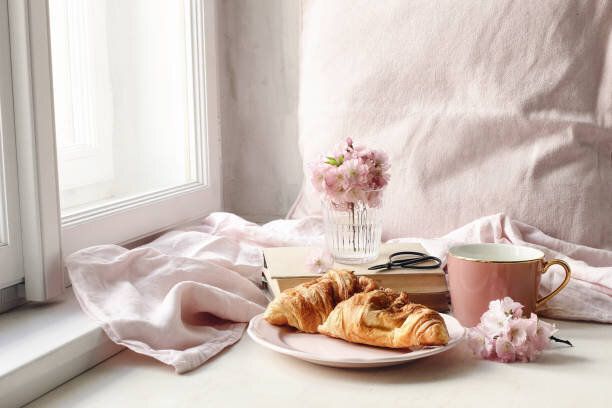 春の静物シーン。コーヒー、クロワッサンペストリー、古い本、はさみのカップ。ヴィンテージフェミニンなスタイルの写真。窓の近くの白いテーブルにピンクの桜、桜の花の組み合い。