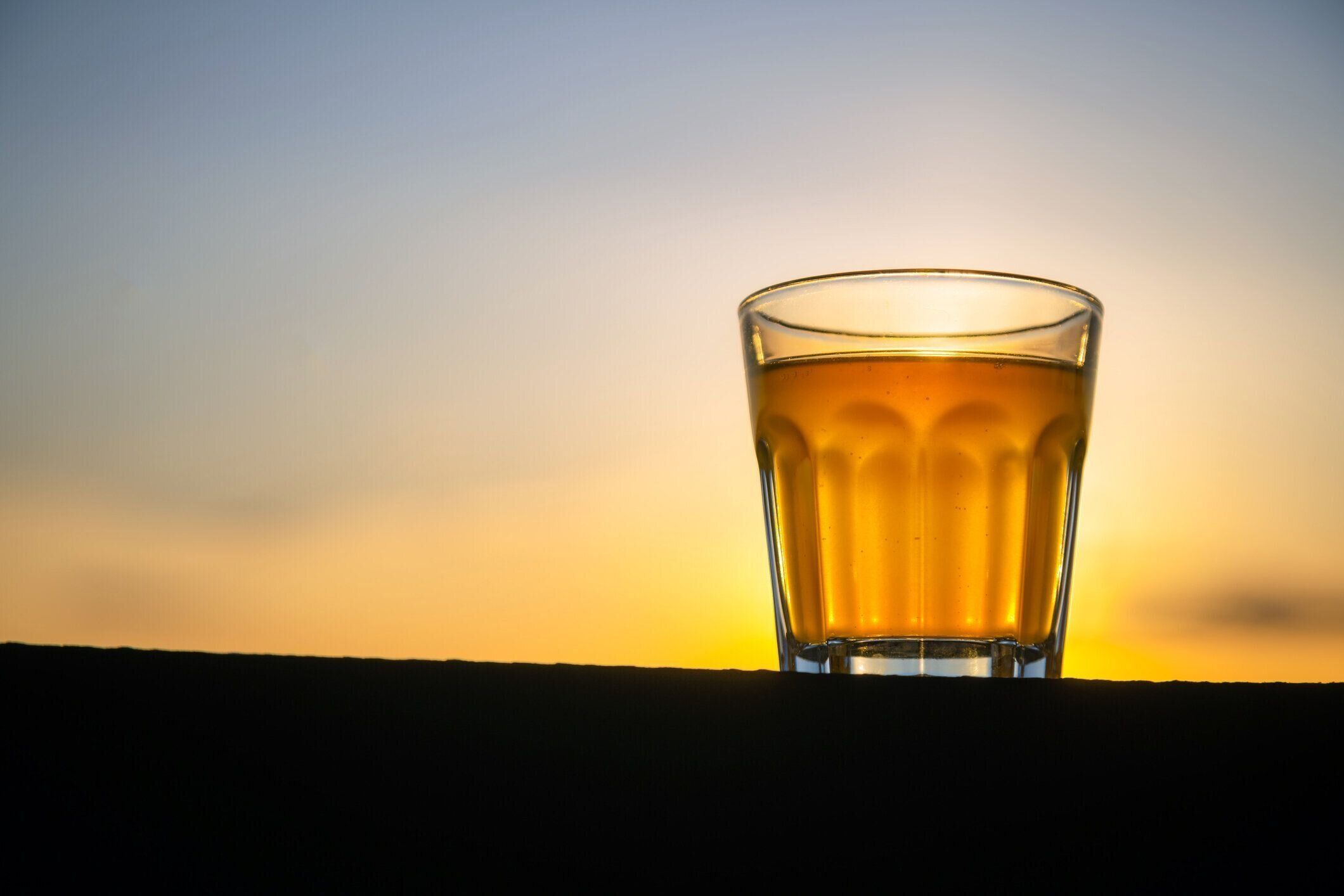 テキストのためのスペースを持つ木製の板の外に泡のないクラフトビールの小さなガラス。日没の背景を持つアルコール飲料のゴールデンサンプル。誰も