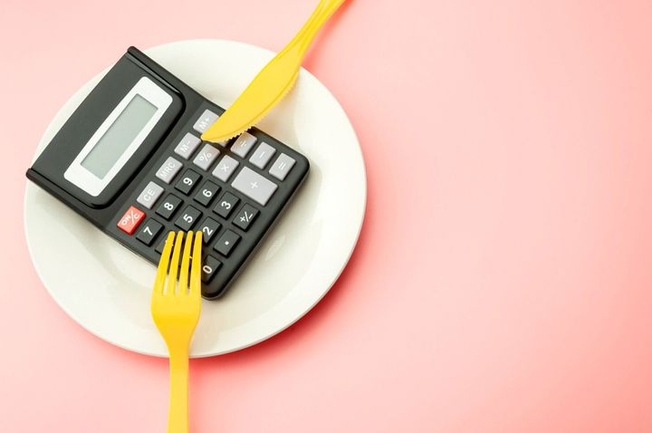 高価な食品支出コストを計算し、空のプレート上の電卓、黄色のフォーク、ナイフをコピースペースでピンクの背景に隔離してカロリーと減量プログラムの概念を数えます