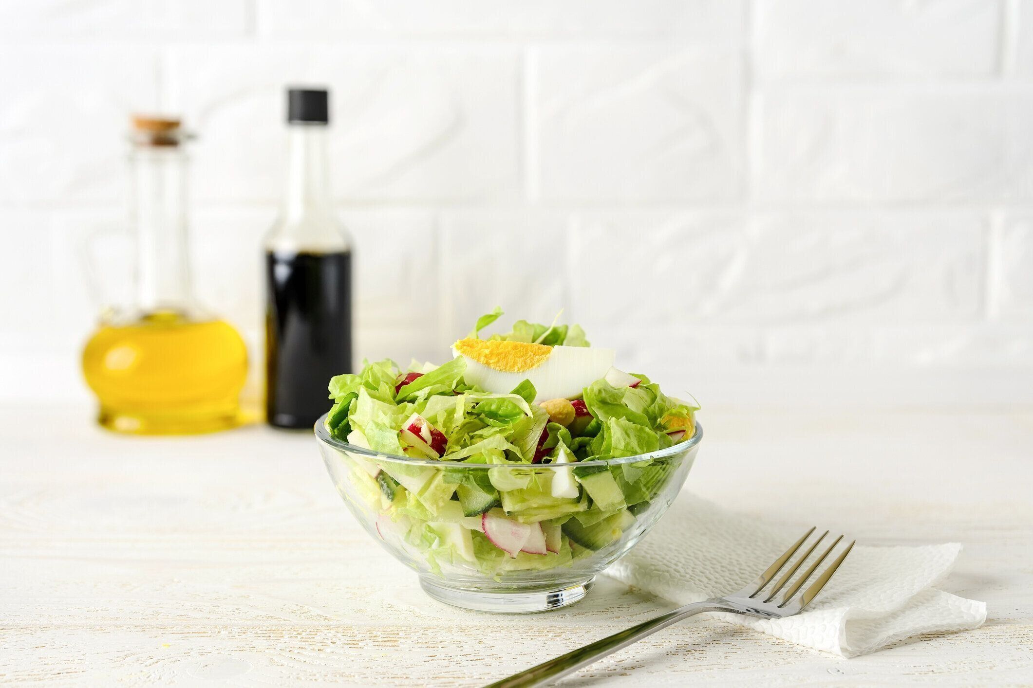 白い木のテーブルの上にガラスボウルにレタス、キュウリ、大根、ゆで卵の新鮮な自家製サラダ。背景にドレッシングのためのオリーブオイルと醤油。健康的な食事。ハイキーショット。