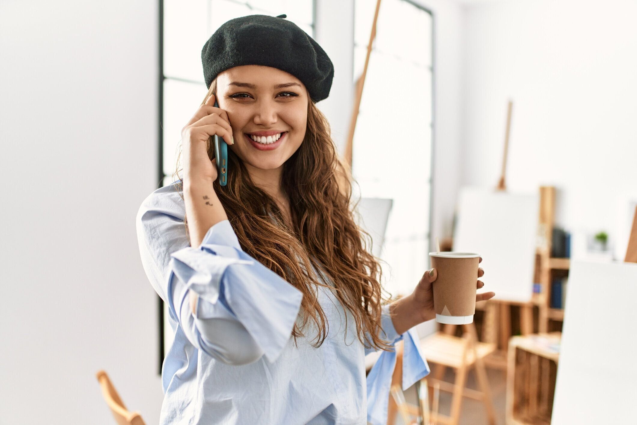 アートスタジオでコーヒーを飲むスマートフォンで話す若い美しいヒスパニック系女性アーティスト
