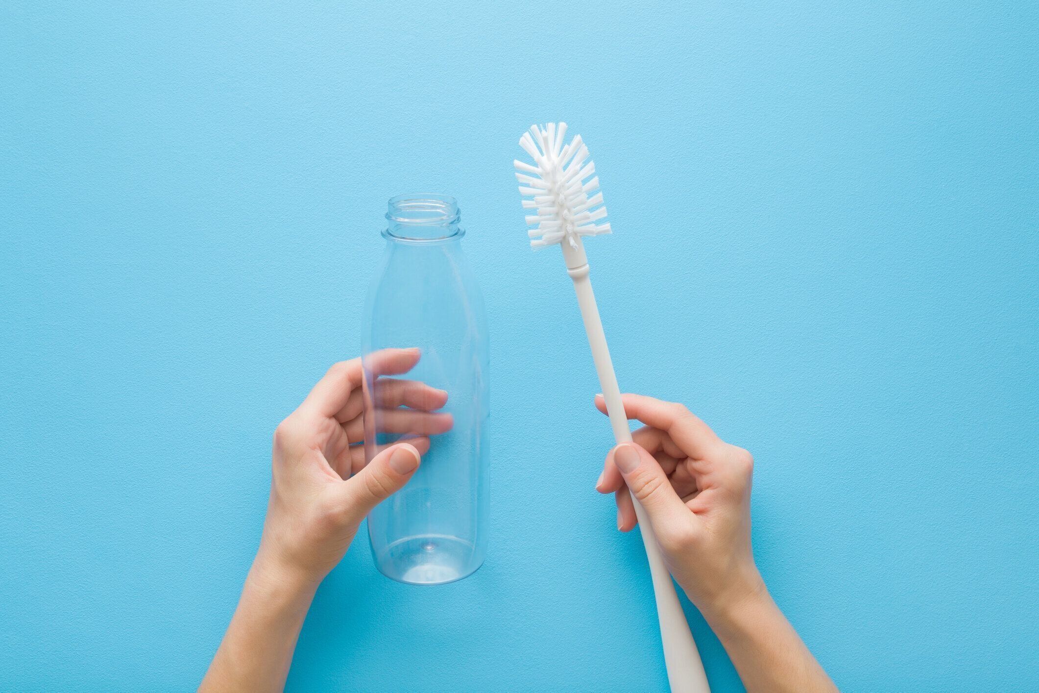 水色のテーブルの背景に白いブラシと透明なペットボトルを持つ若い大人の女性の手。パステルカラー。視点ショット。クローズ アップ。上から見下ろしたビュー。