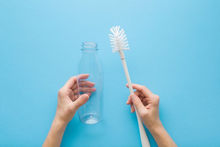 水色のテーブルの背景に白いブラシと透明なペットボトルを持つ若い大人の女性の手。パステルカラー。視点ショット。クローズ アップ。上から見下ろしたビュー。