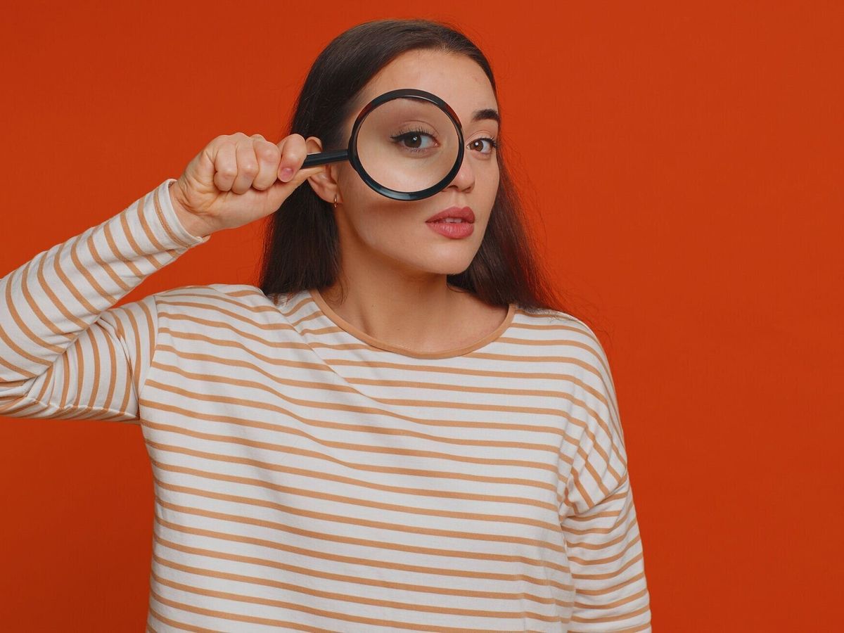 顔の近くに虫眼鏡を持った調査員研究員の女性、大きなズーム目でカメラを覗き込む