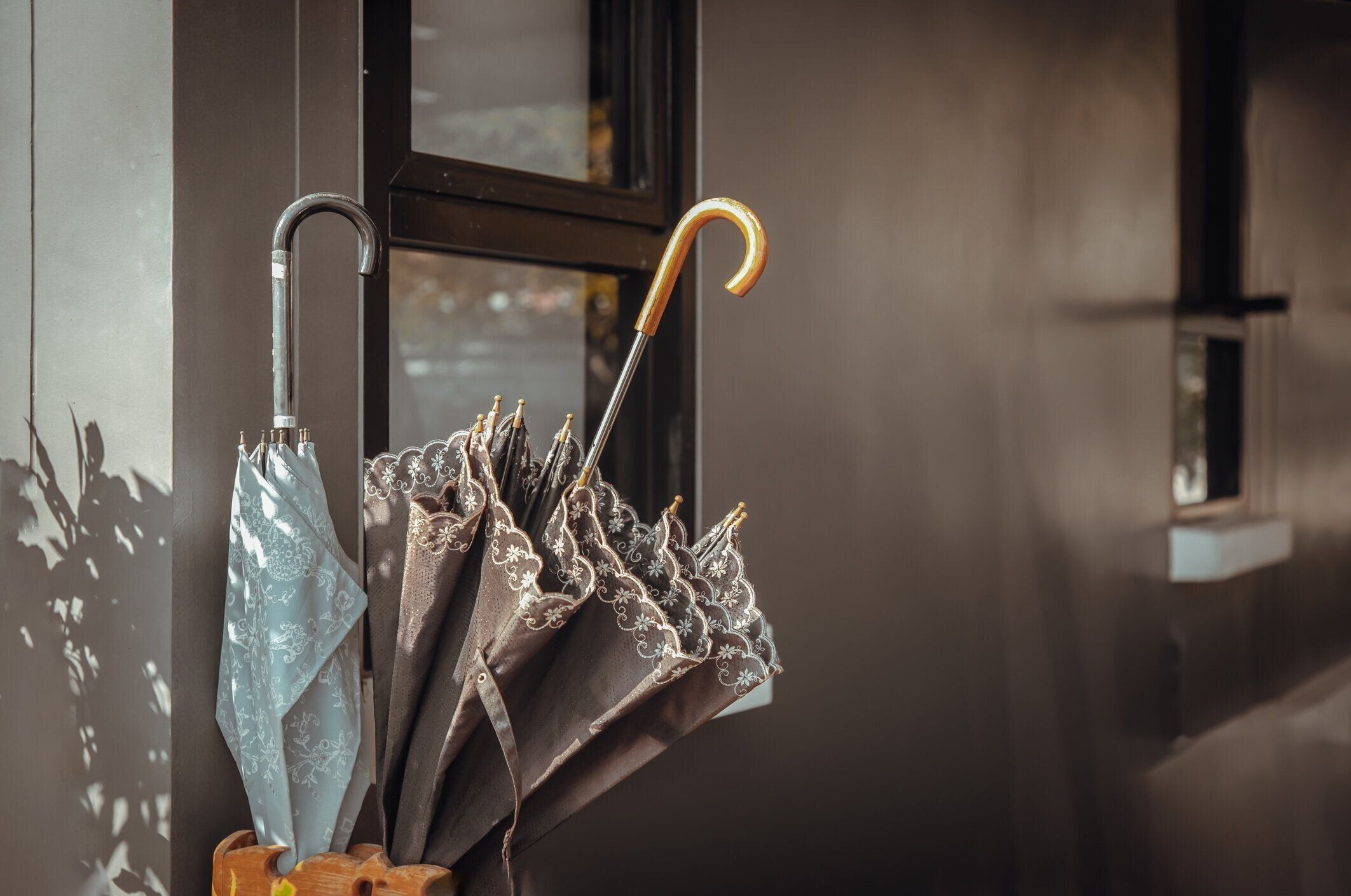 家の外側壁の脇にある木で作られた傘置き場の茶色と青の傘に太陽の光が差し込みます。