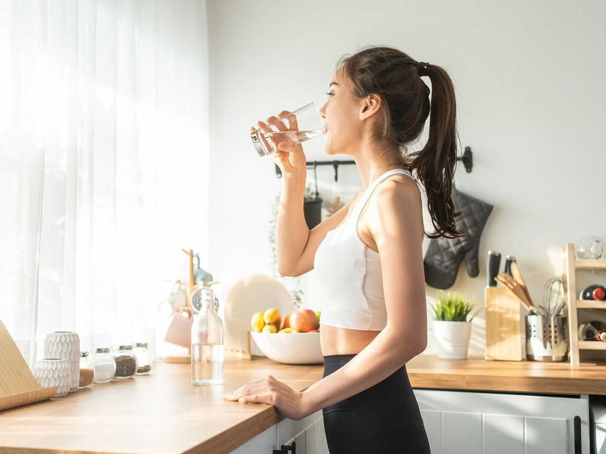 スポーツウェアを着たアジアの美しい女性は、自宅で運動した後に水を飲みます。若いのどが渇いたアクティブスポーツの女の子は、家の台所でヘルスケアのためのトレーニングの後、カッ�