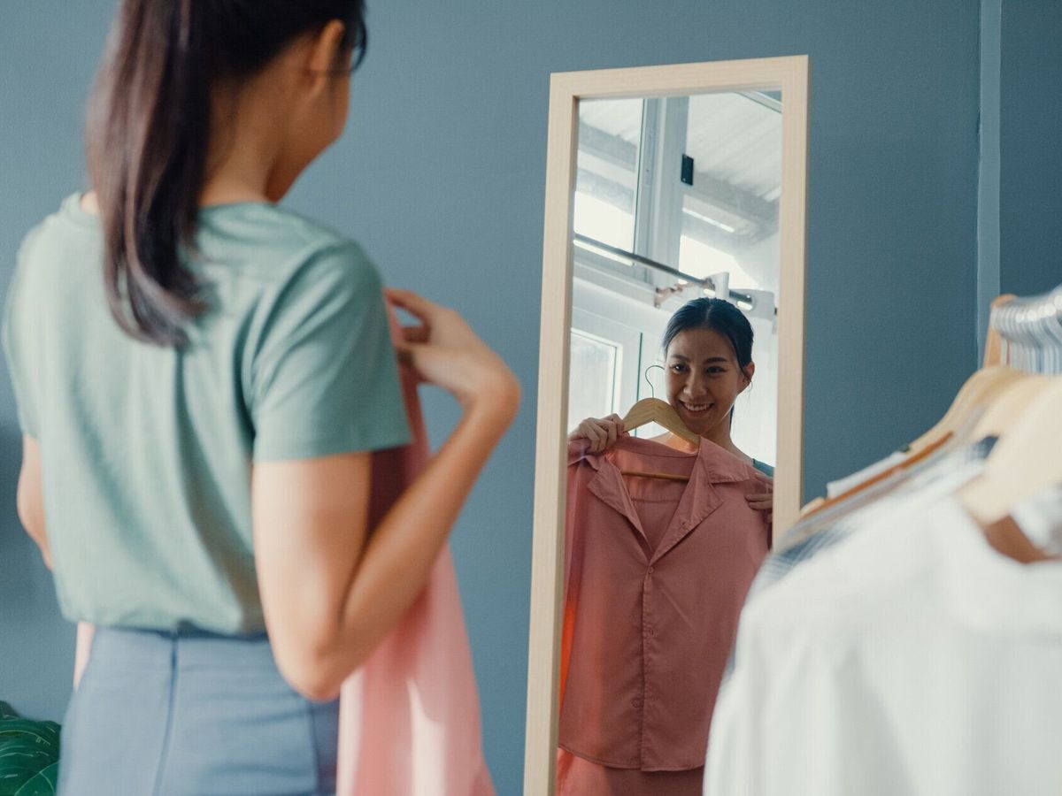 家のリビングルームで鏡で自分自身を見て服ラックドレッシングに服を選ぶ美しい魅力的なアジアの女性。