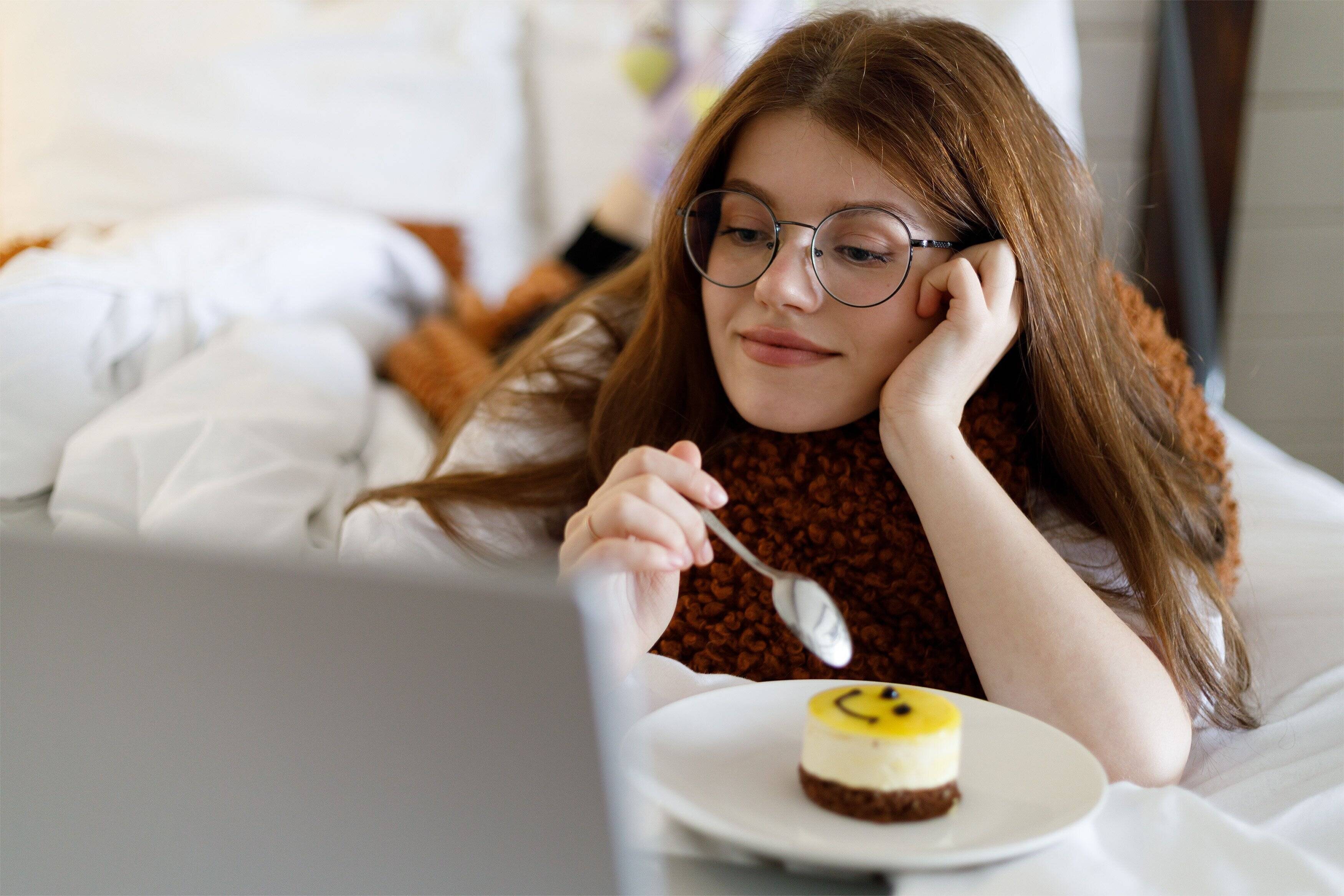 美しい若い女性がラップトップを持ってベッドに横たわり、映画を見たり、お菓子を食べたりしています。