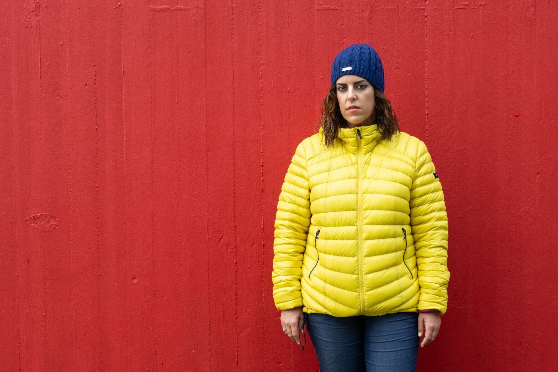 黄色のアノラックと青いウールの帽子をかぶった30代のブルネットの白人女性の肖像画は、彼女の後ろに深い赤い壁でポーズをとっています