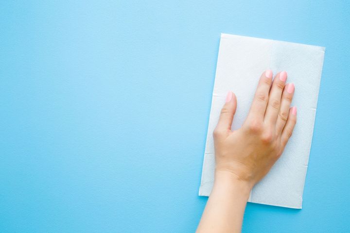 女性の手は白い紙ナプキンでパステルブルーの机を拭きます。一般的または定期的なクリーンアップ。閉じる。テキストやロゴのための空の場所。トップビュー。