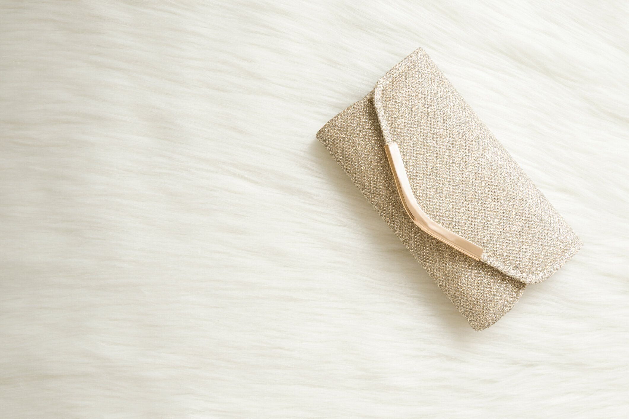 白いふわふわの毛皮の背景に光沢のある、黄金のクラッチバッグ。女性アクセサリー。テキストの空の場所。クローズ アップ。トップダウンビュー。