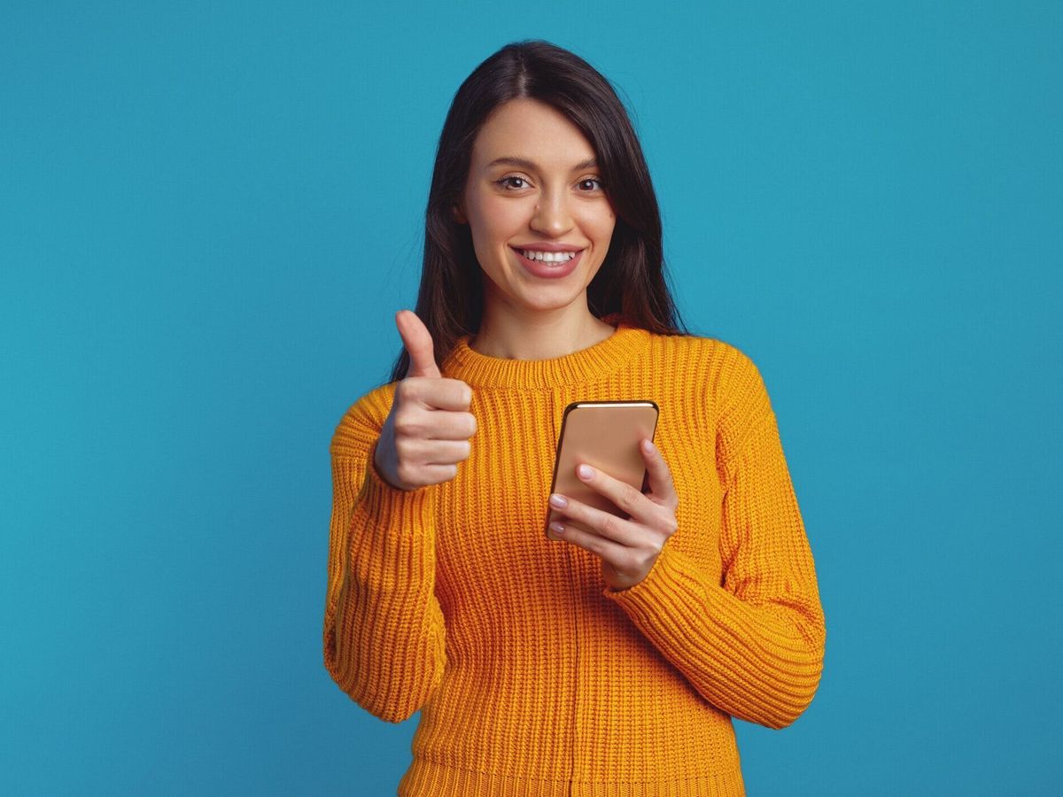 スマートフォンを使用しながら親指を表示するオレンジ色のセーターを着た若い美しい女性