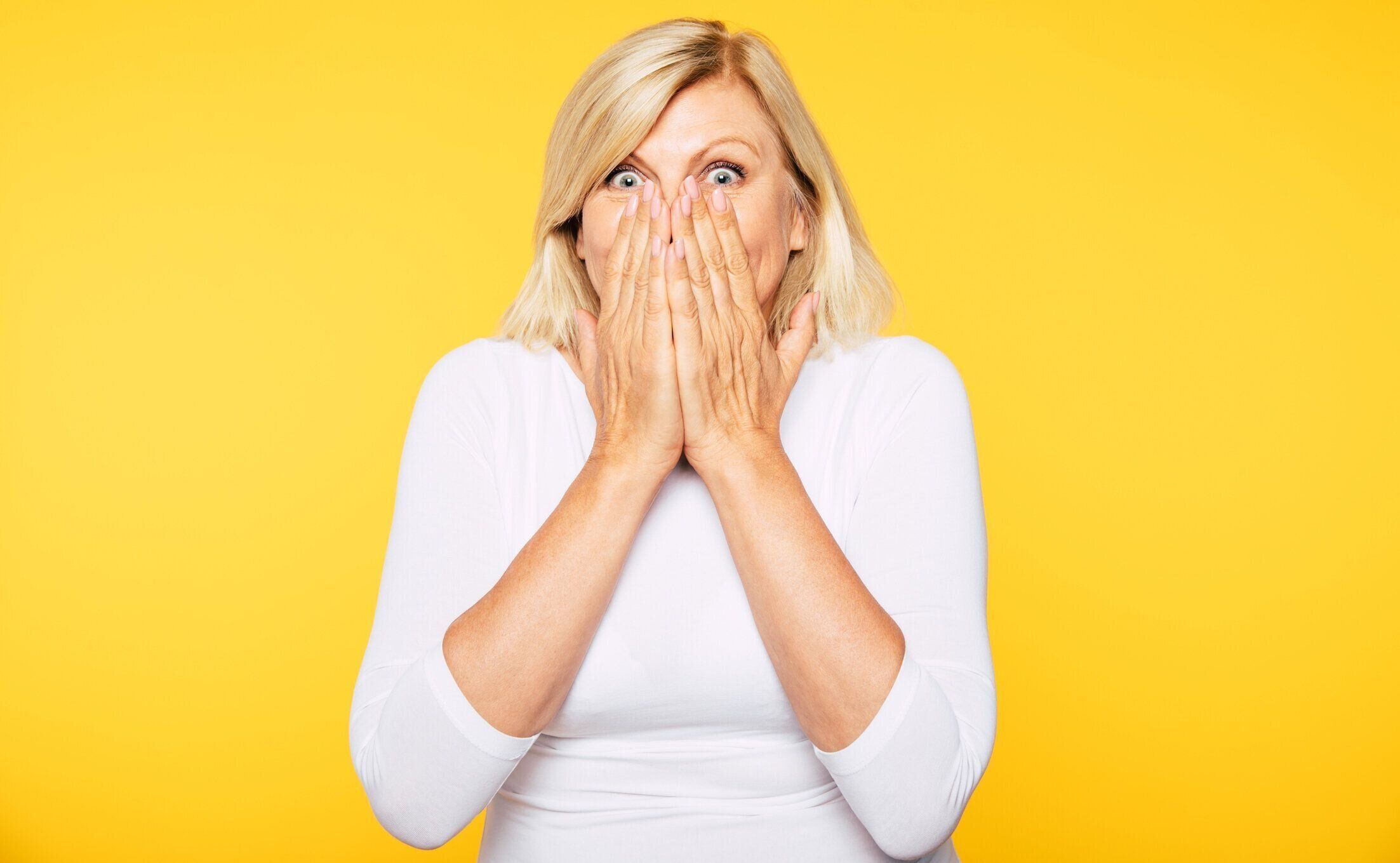 話さないで下さい！ブロンドのシニア女性は手の助けを借りて彼女の口をカバーし、目にショックでカメラを見ます。