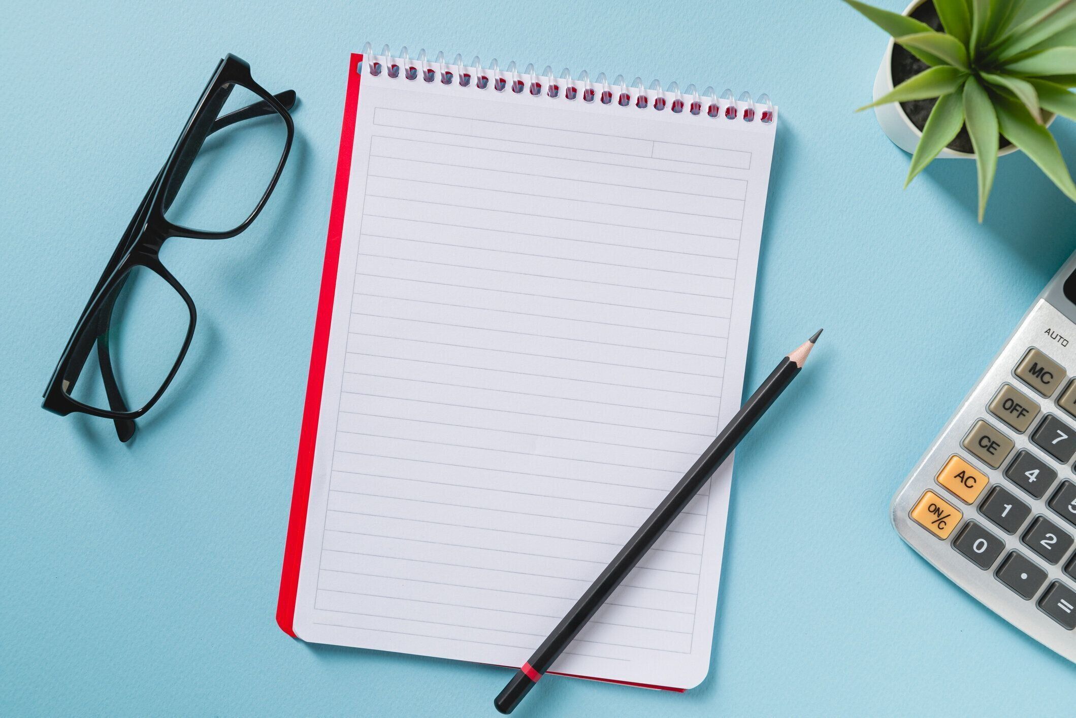 青いオフィスの机の上に空白のノートブック、メガネ、ペン、電卓、緑の植物の上から見た図