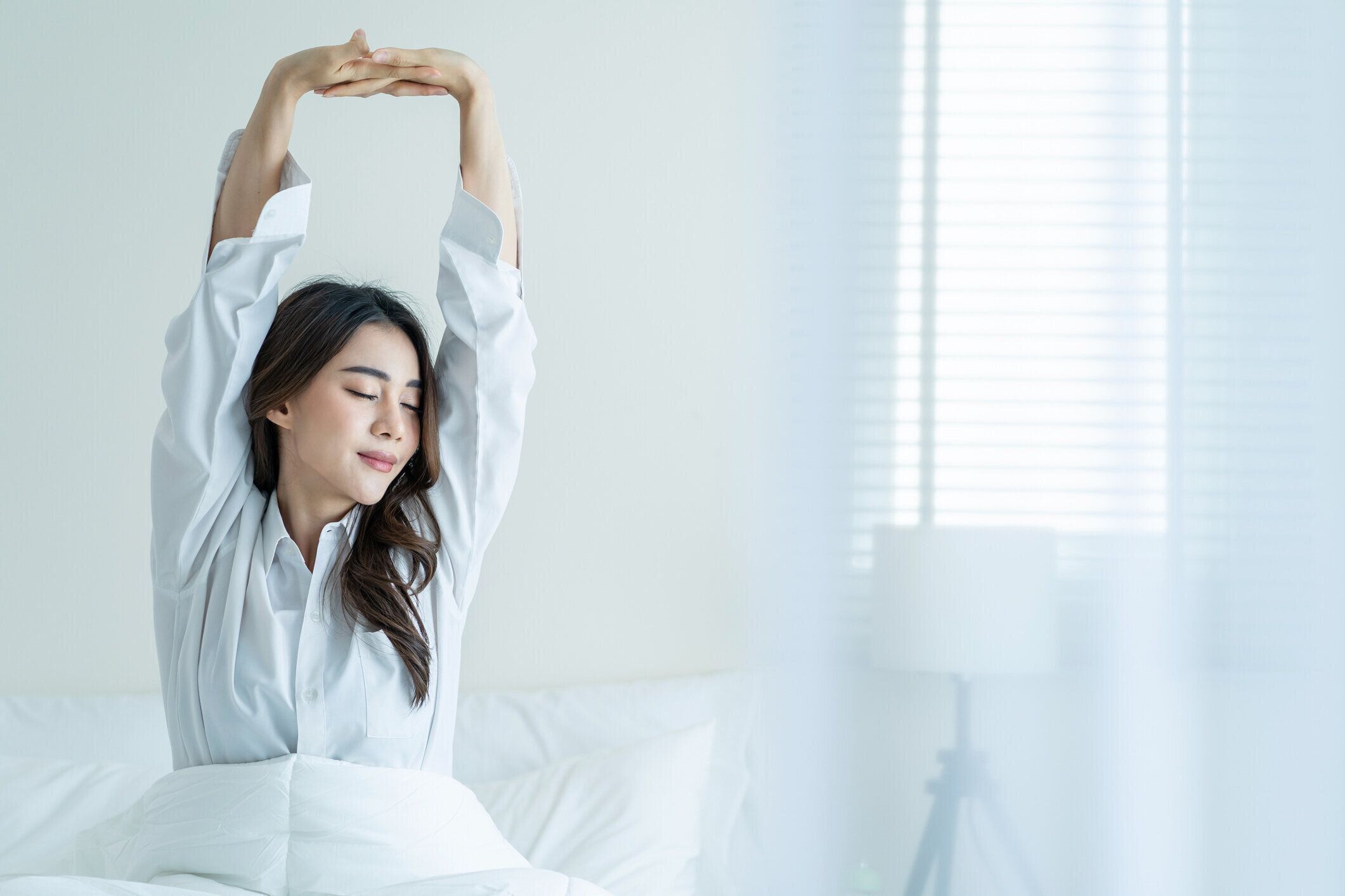 パジャマを着たアジアの美少女が朝、幸せで目を覚ます。魅力的な若い女性は笑顔で、幸せを感じ、自宅の寝室でベッドで寝てから起きた後、体を伸ばしてリラックスしてください。