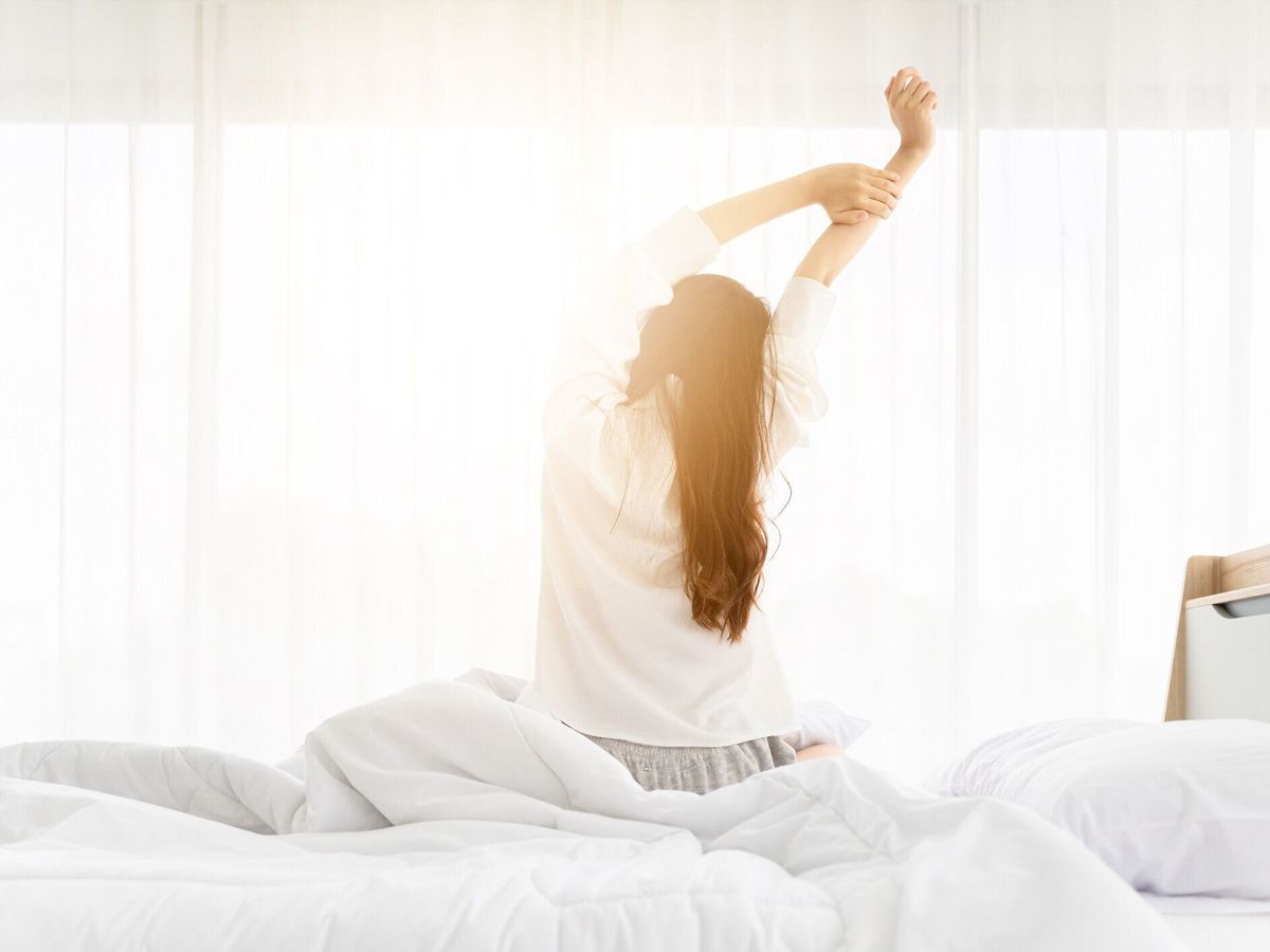 おはようございます新しい日。アジアの女性が目を覚ますと、寝室の窓の横のベッドに座って体を伸ばす