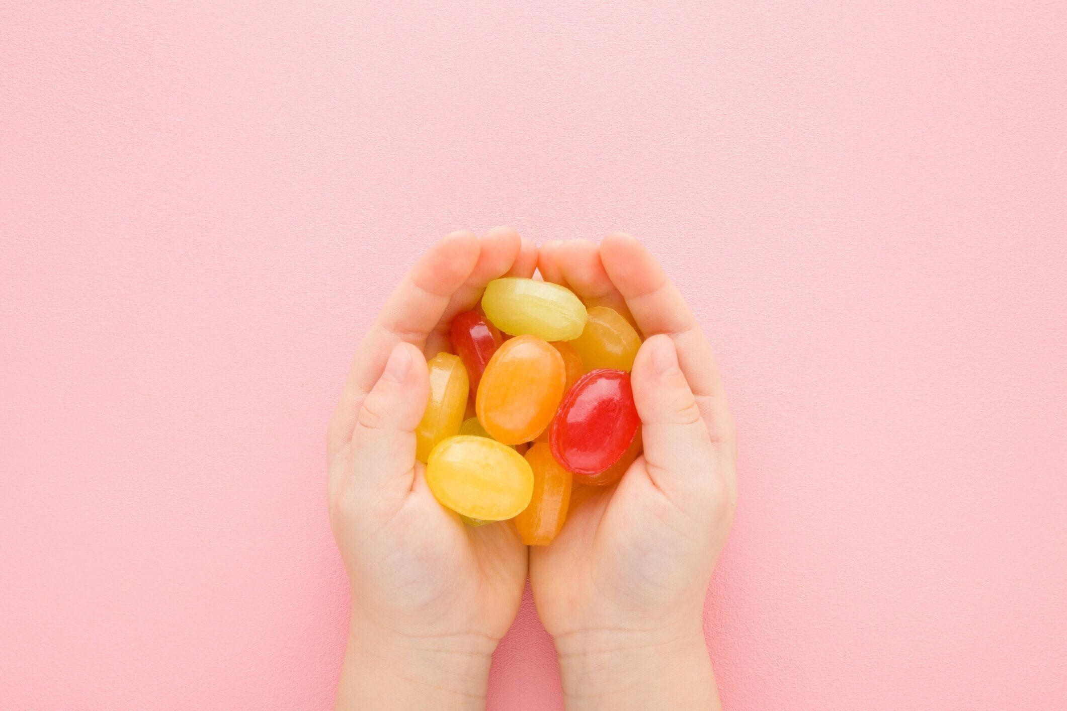 明るいピンクのテーブルの背景に女の赤ちゃんのカラフルな砂糖菓子が手のひらを開いた。パステルカラー。甘いおやつ。クローズ アップ。視点ショット。トップダウンビュー。