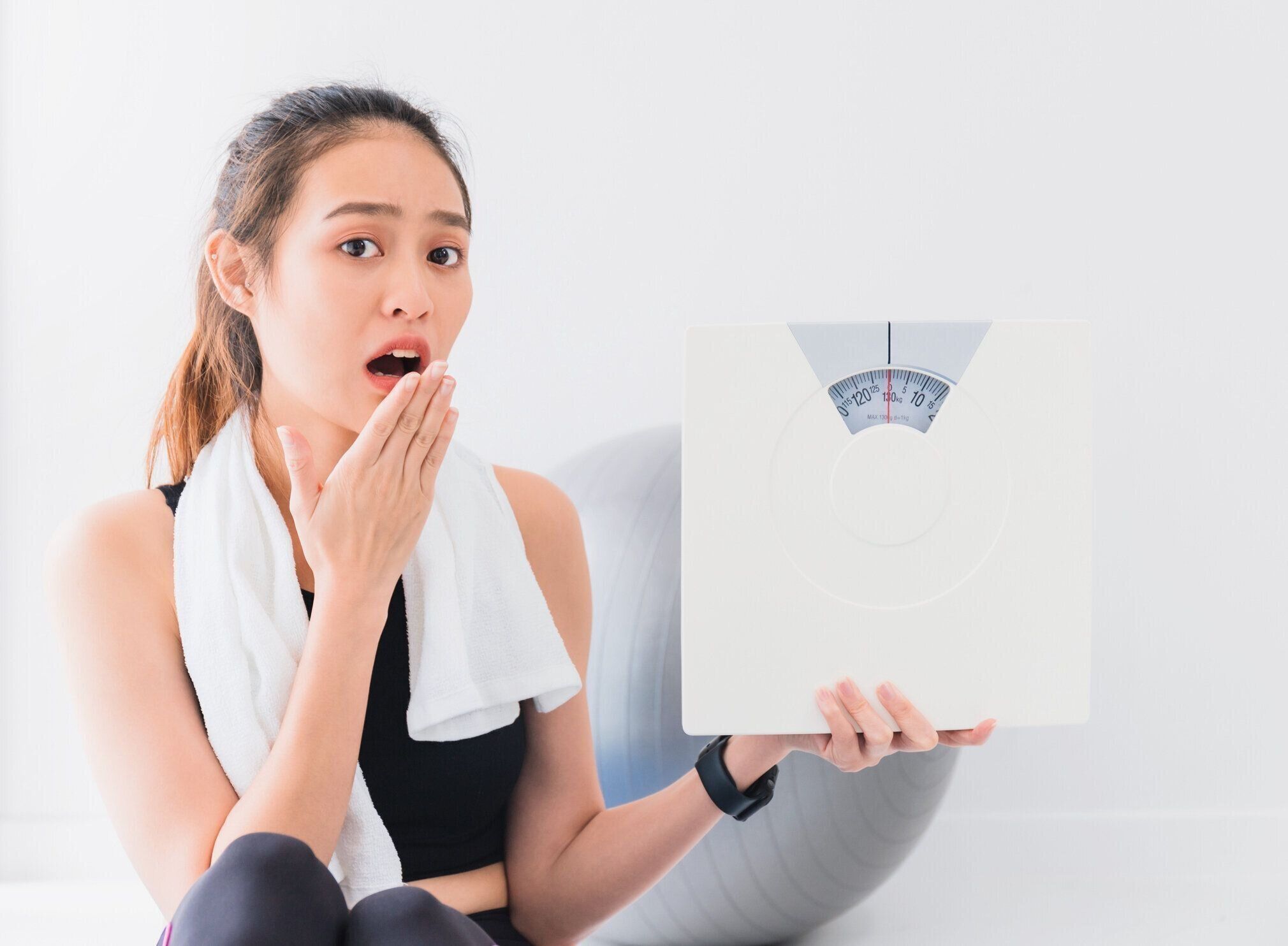アジアの女性は、自宅でヨガや運動をした後、驚き、体重計を保持しています。体重、減量についての心配を表現します。