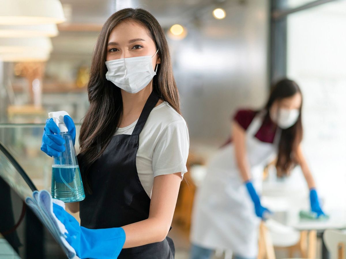 朝の安全で清潔なカフェを消毒する際に柔らかい布でそれを拭くカウンター製品キャビネットに均一なエプロンスプレー洗剤やアルコールを身に着けた魅力的な肯定的な若いアジアの女性ス�