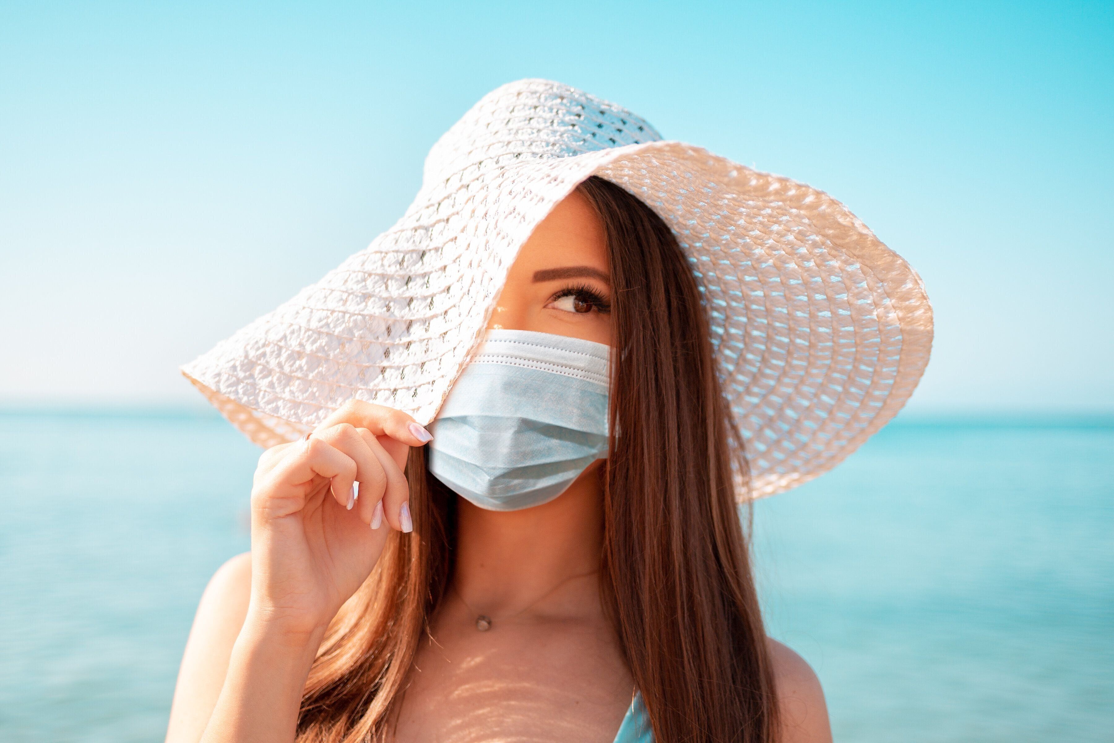 麦わら帽子と保護マスクを着た若い女性の肖像画は、思慮深く帽子の端で彼女の顔を覆っています。背景の海。コロナウイルス中の休暇の概念