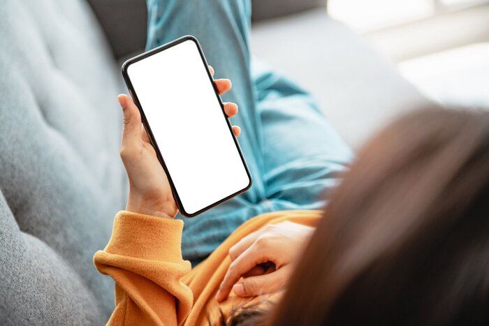 リビングルームのソファに空白の白い画面を持つ携帯電話のスマートフォンを使用している女性。