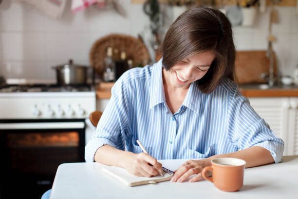 紙のノートにメモを書く若い女性。コーヒーのカップとテーブルに座って幸せな女の子