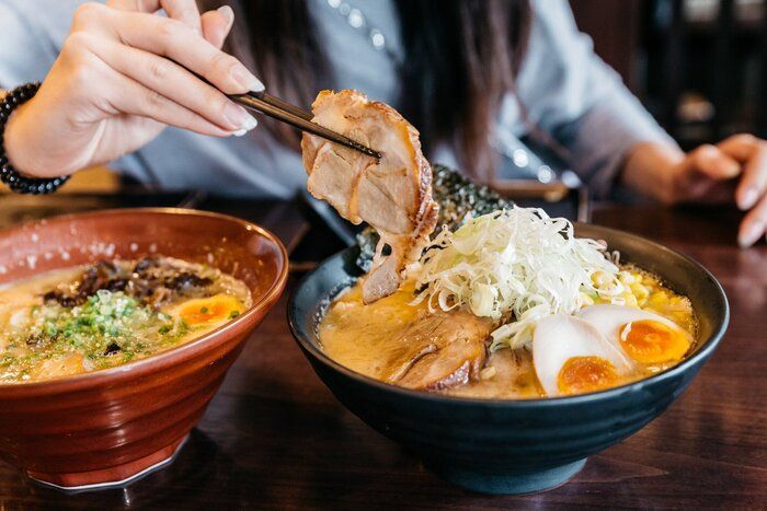 チャーシュー豚、葱、もやし、とうもろこし、干し海藻、ゆで卵を添えたラーメン豚骨スープ (とんこつラーメン) で手をつまんでいる女性がブラックボウルでお召し上がりいただけます。